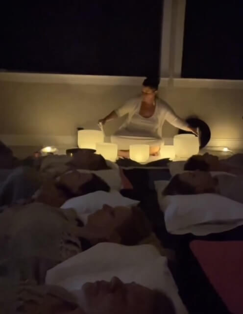 A group of people lying down on mats during a candlelit sound bath meditation session with a practitioner playing crystal singing bowls.