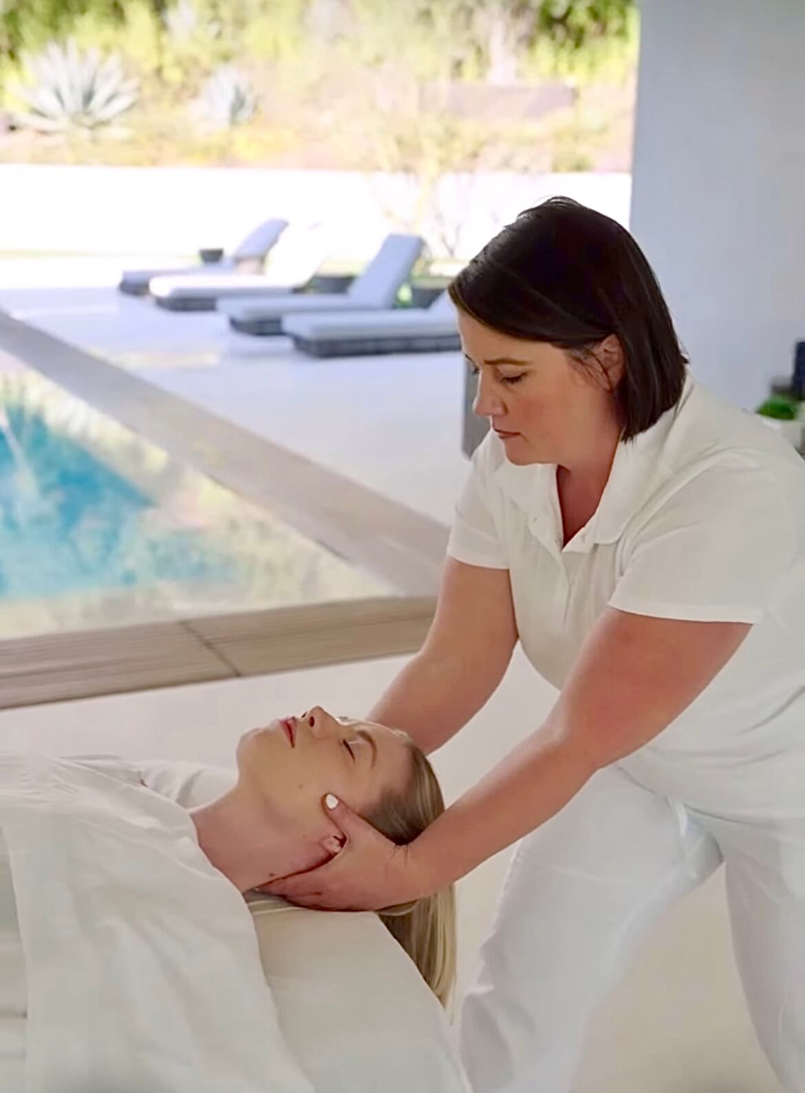 A massage therapist performing cranial sacral head therapy on a client poolside at a luxury modern resort.