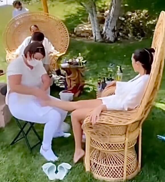 A client in a white robe relaxing in a large wicker peacock chair during an outdoor foot reflexology treatment in a garden setting.
