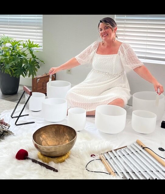 A smiling wellness practitioner in a white dress sitting on a rug surrounded by a variety of white crystal singing bowls and sound healing instruments.